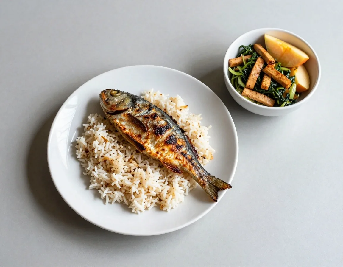 Simple home-cooked meal with rice, grilled fish, greens and tempeh on a pale table